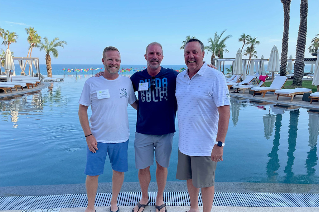 Three men standing in front of a pool with palm trees.