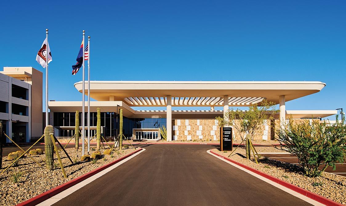 Front view of the Gila River Santan Mountain Casino with a unique, wide white canopy entrance, desert landscaping, and American and state flags flying under clear blue skies.