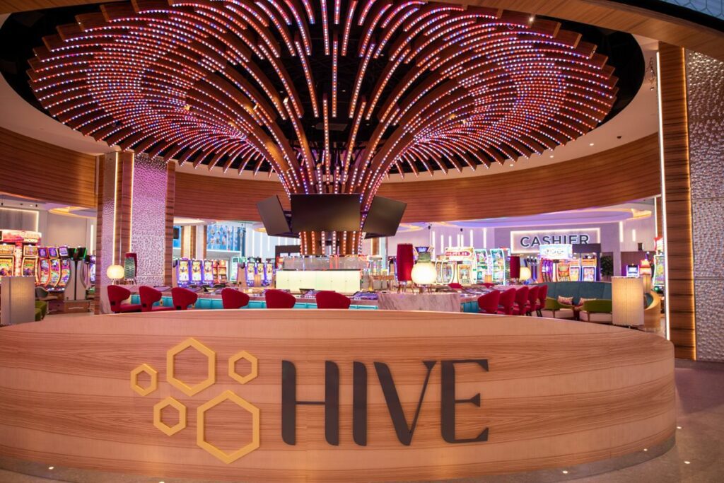 A modern casino lobby with a bar named "hive" under a vibrant, honeycomb-patterned LED ceiling at the Gila River Santan Mountain Casino, surrounded by slot machines and a cashier's