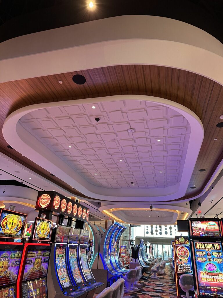 Modern casino interior at Gila River Santan Mountain Casino with colorful slot machines lined up near large windows, featuring an elegant vaulted ceiling with wooden accents and white panels.