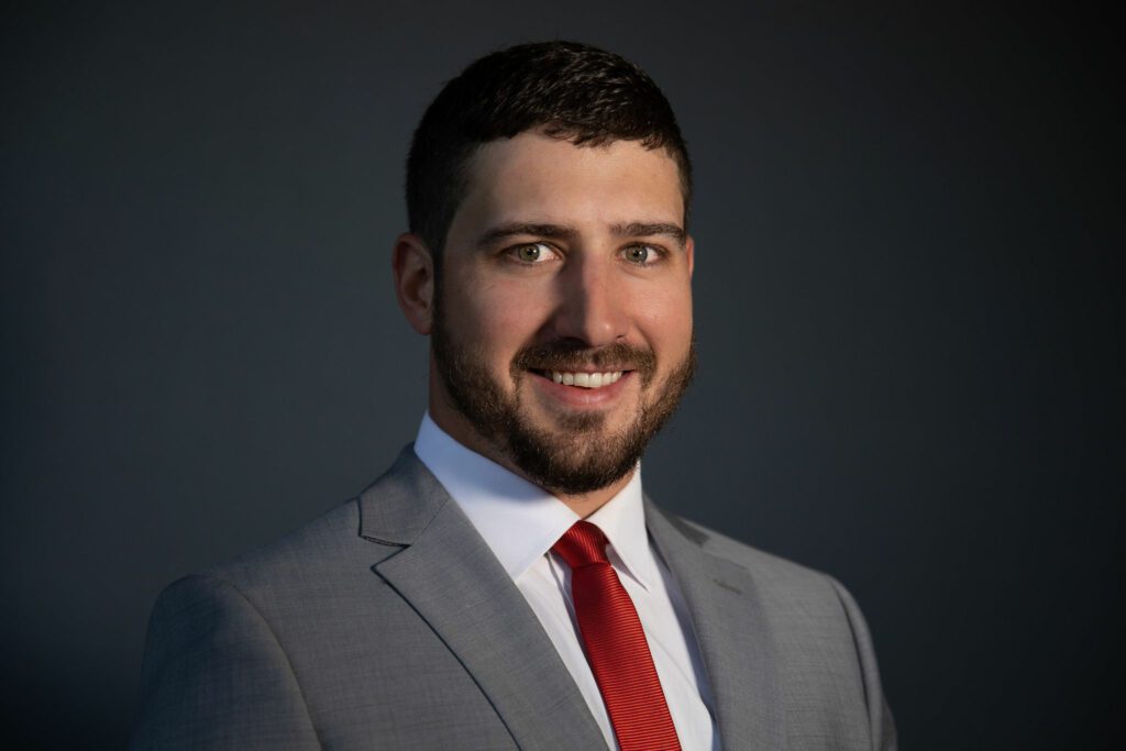 A professional portrait of a man with a beard, wearing a gray suit, white shirt, and red tie, smiling against a dark background for MKB Construction Inc.