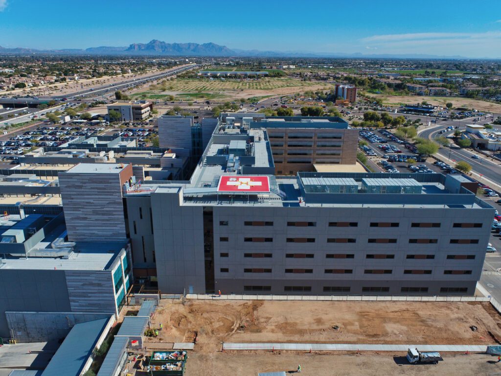 Aerial view of a large hospital complex, including the Banner MD Anderson Cancer Center, with connected buildings, parking lots, and roads, with mountains in the background on a clear day.